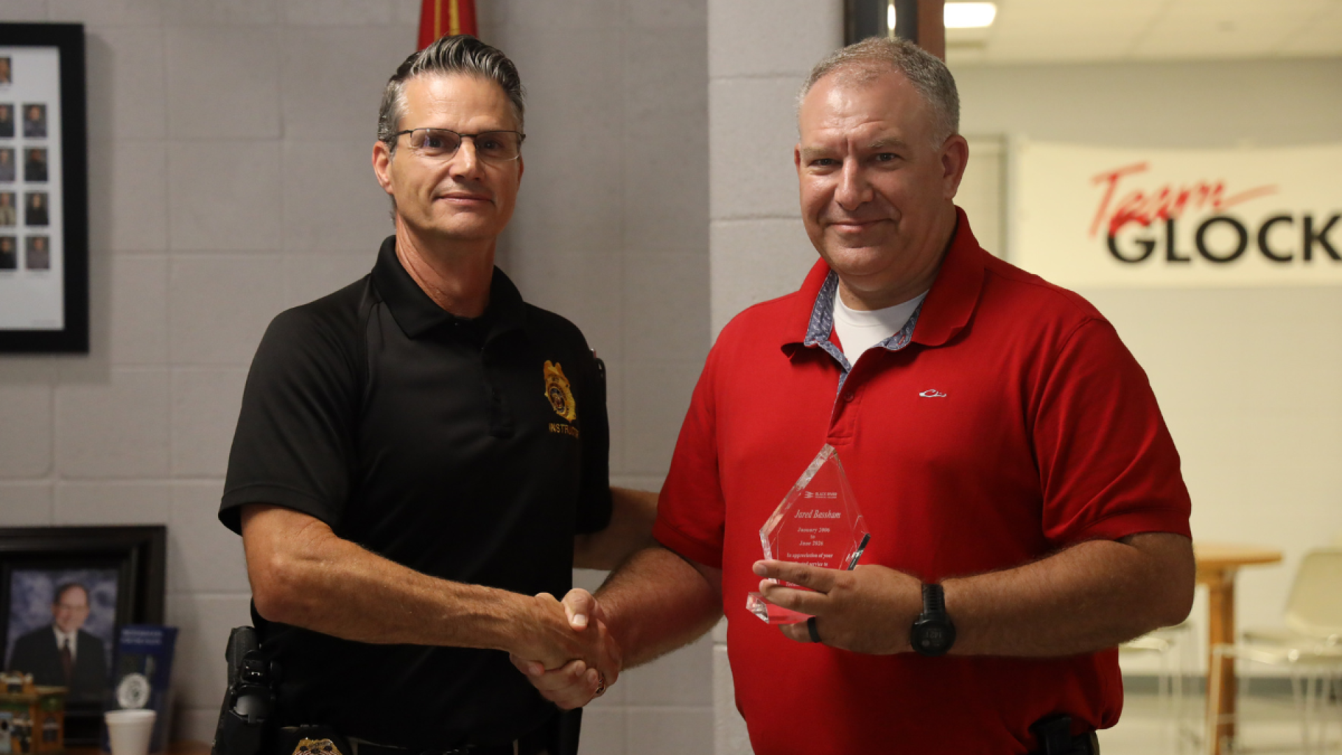 A man in a black instructor polo shakes hands with a man in a red polo holding a glass award.